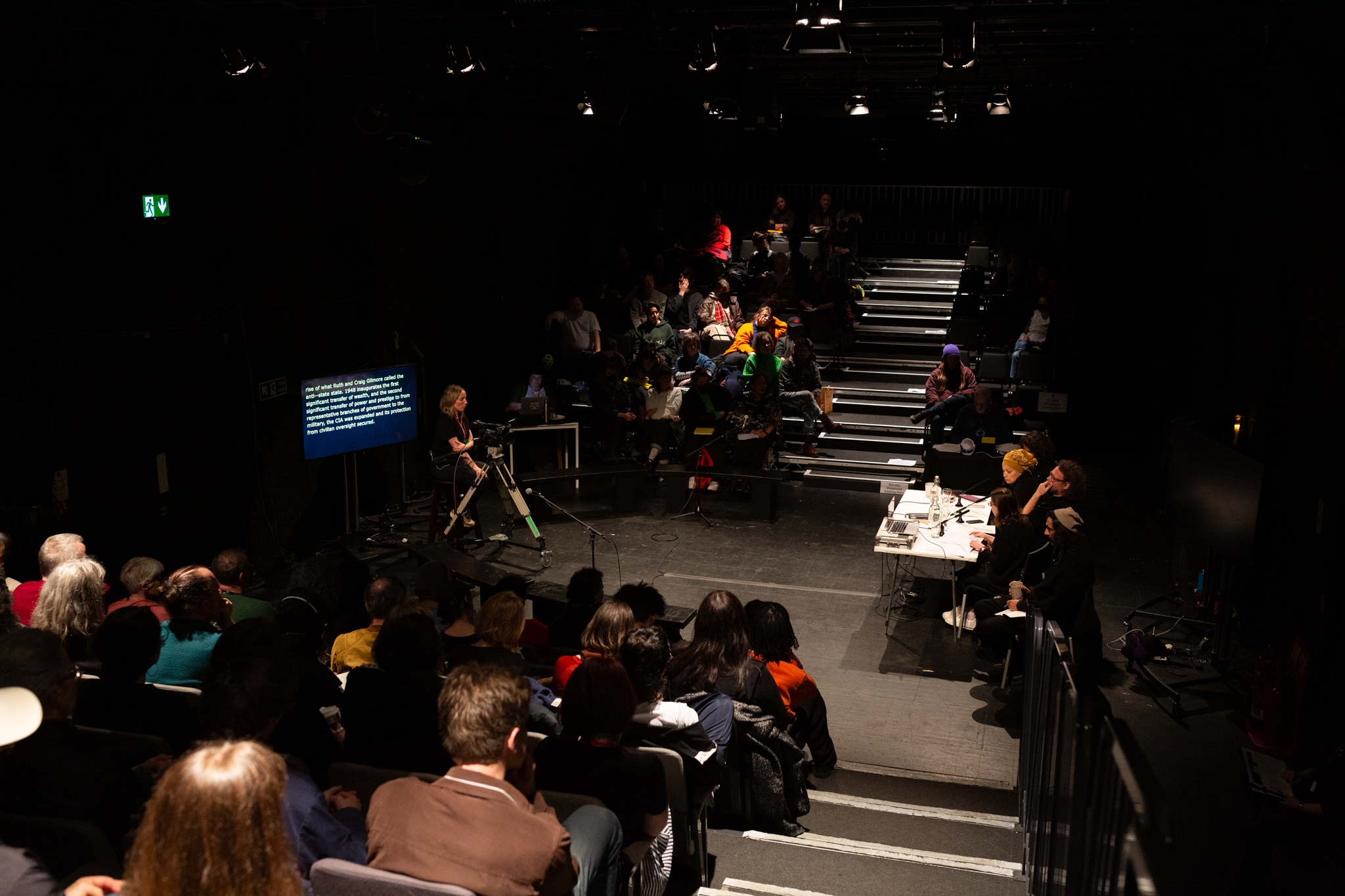 A theatre space in the round. In the middle is a table with people sat at a desk, to either side are seating banks filled with audience. Infront of the speakers is a person operating a camera and being them is a screen with live captioned text