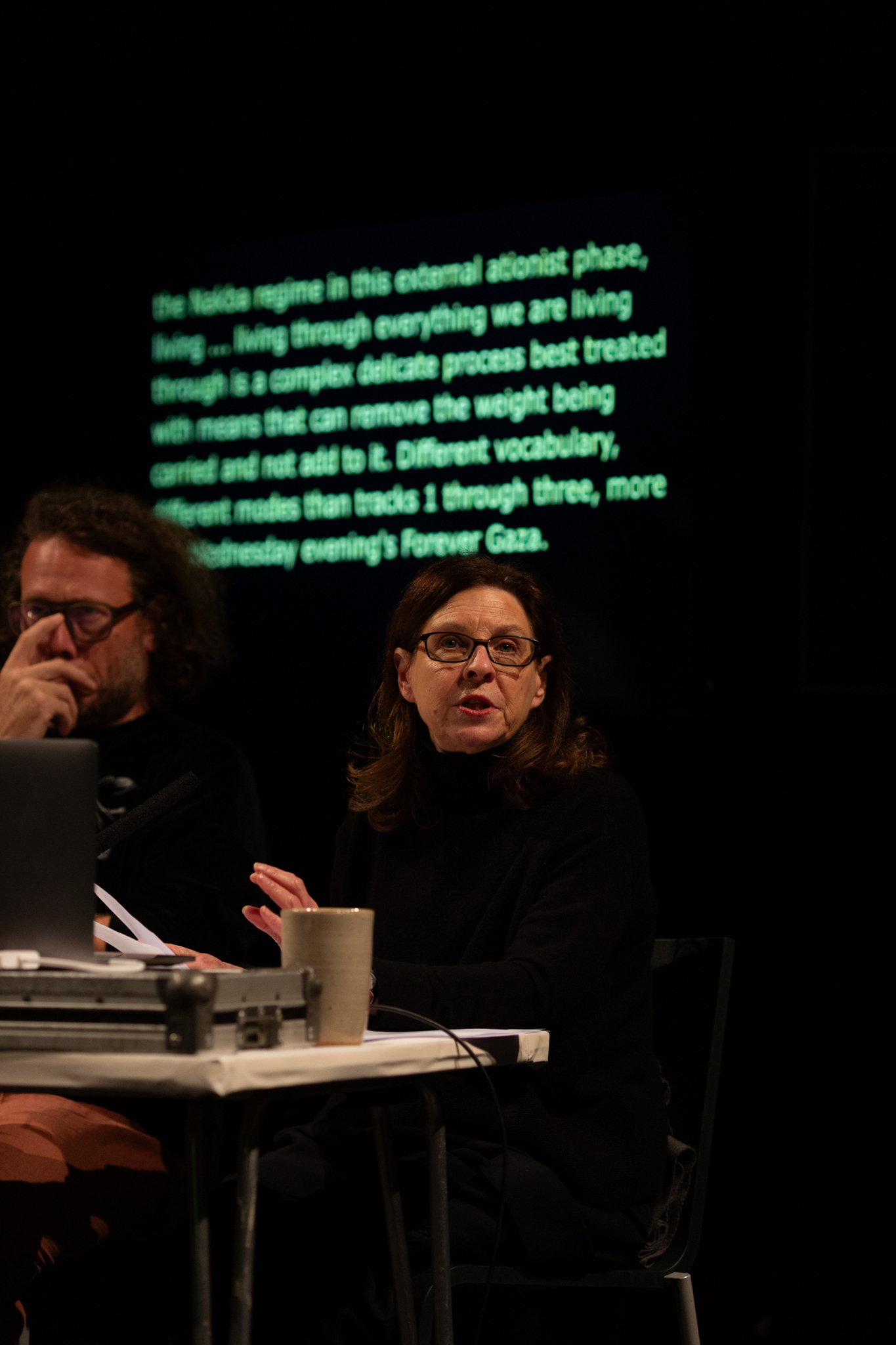 A person sat at a desk talking while gesturing with their hands. They are sat at a white desk, behind them is a screen with green glowing text but it is slightly blurred and hard to read.