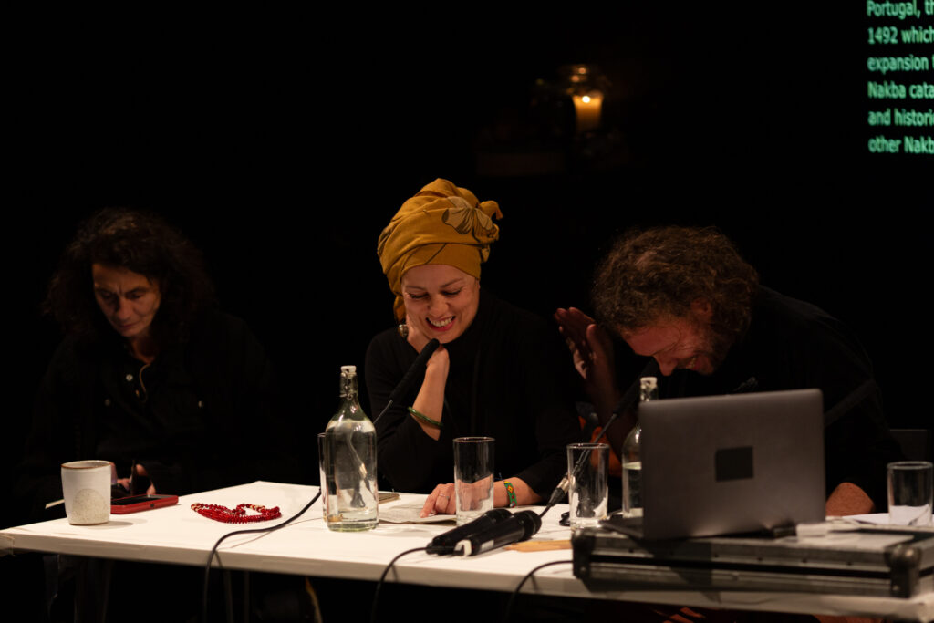 Two people are sat smiling and laughing while looking downwards. One is about to put their hand on the others shoulder. They are sat at a white desk which has microphones, drinks and. laptop on it