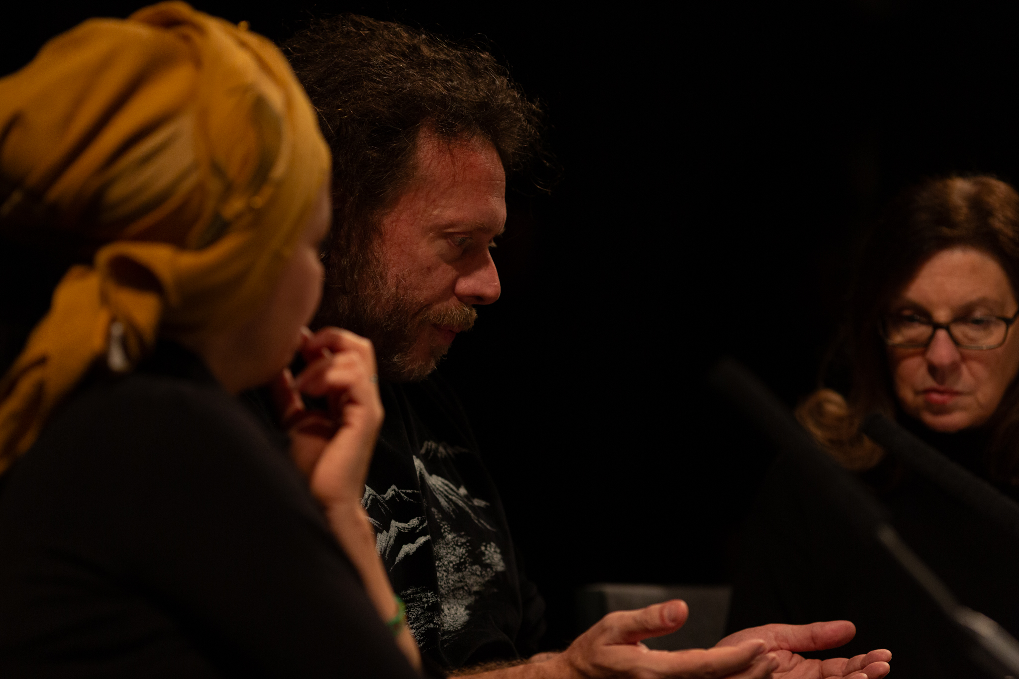 Someone sitting, head tilted slightly downwards with their hands open in front of the them with the palms up. Two people sit at either side, one looks downwards and the other we see the back of their head which is covered in a yellow head scarf