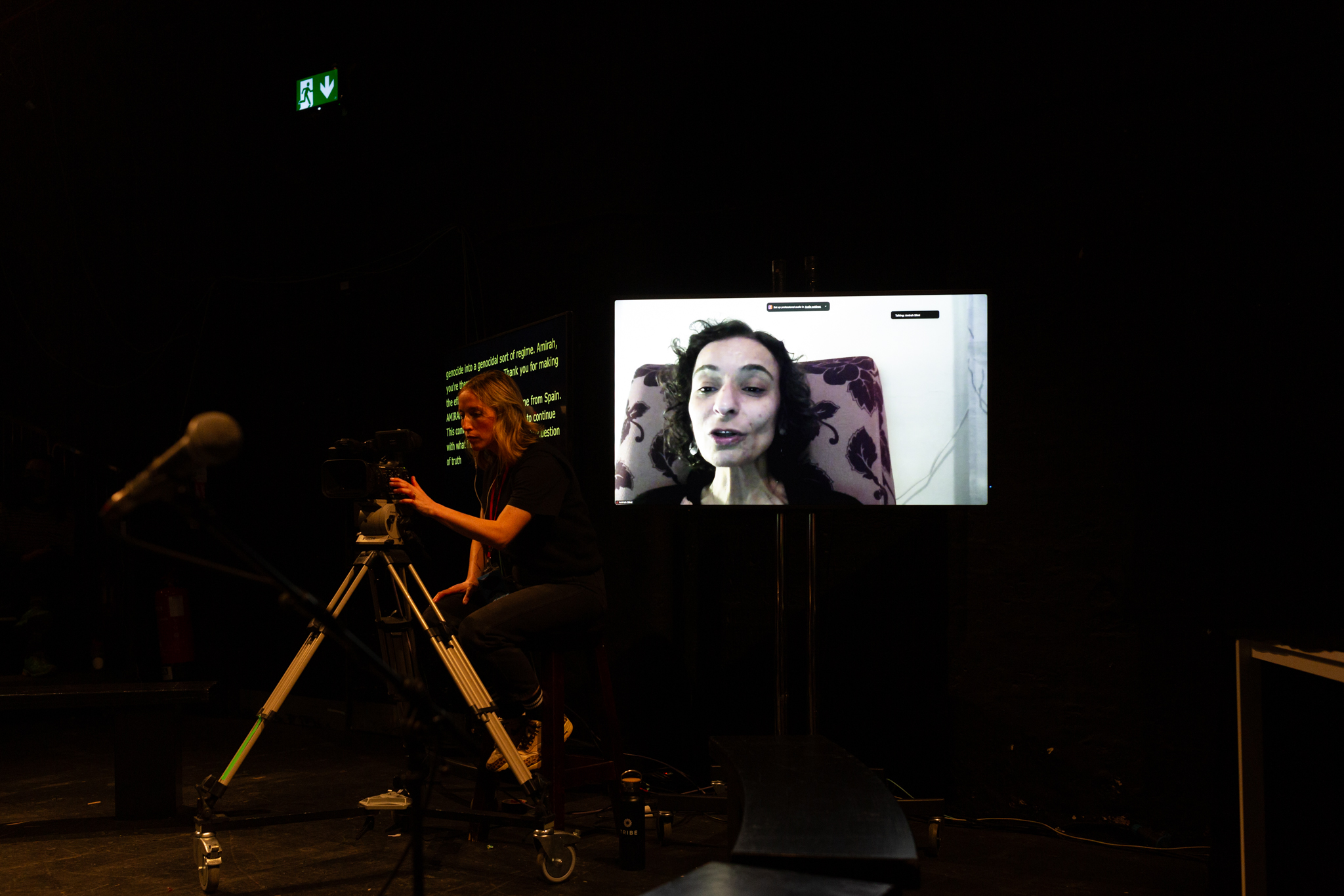 A screen from a video call with a person talking sat on a floral chair. Next to the screen someone stands operating a camera and behind them is a second screen with yellow captioned text