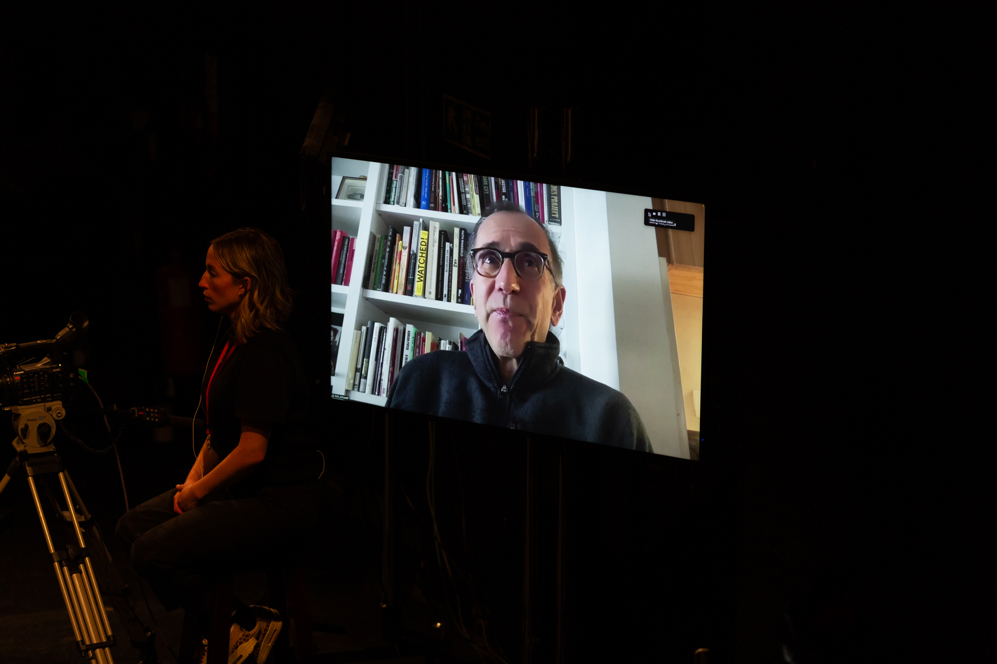 A screen from a video call with a person sat infront of a white bookcase filled with books. Next to the screen someone stands operating a camera