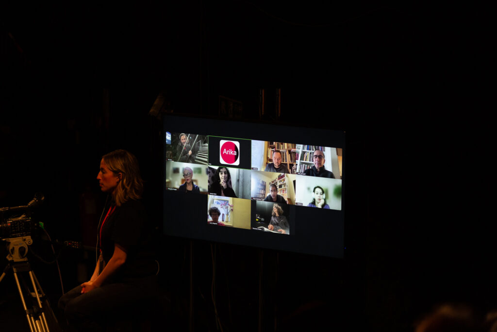 A screen from a video call with several participants. To the front of the screen someone stands operating at camera