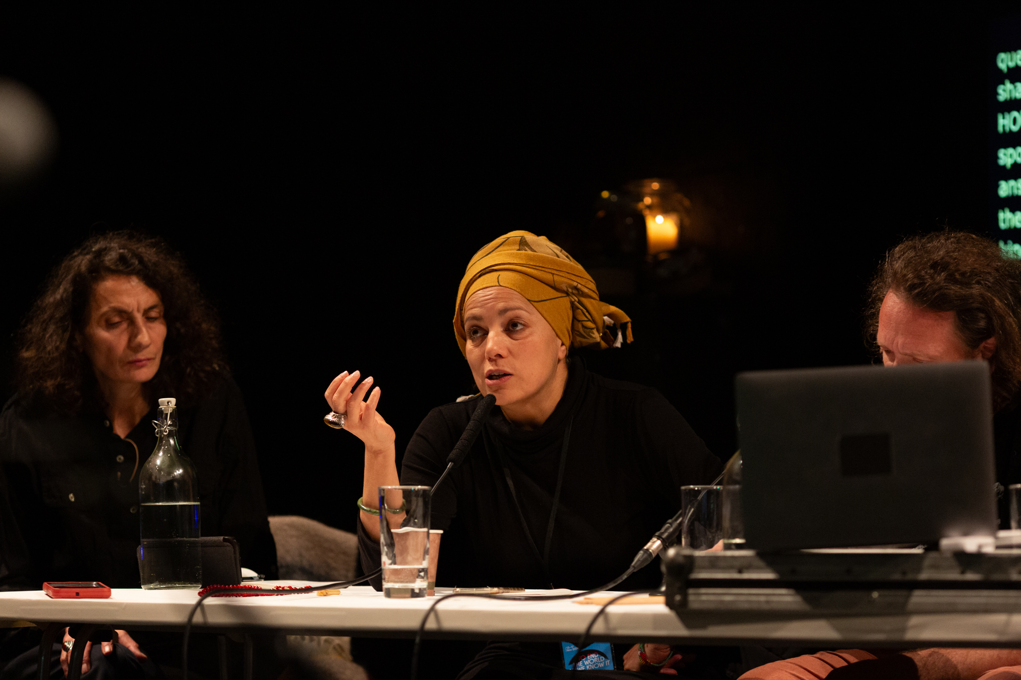 A person wearing a yellow head scarf talks into a microphone that sits on a white table which has drinks and a laptop on it. The background it dark with a glowing candle in the background. Two other people sit at the table listening