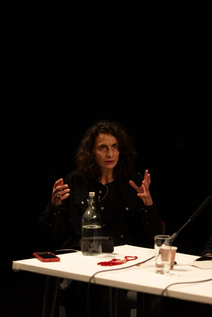 A person talking expressively gesturing with their hands. They are sat at a white table which has drinks, microphones & a phone on it. The background is dark