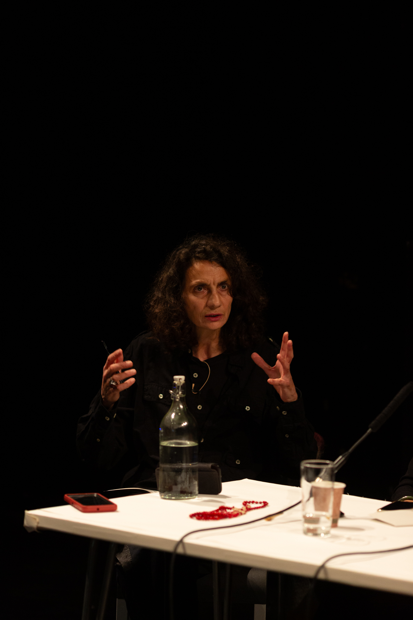 A person talking expressively gesturing with their hands. They are sat at a white table which has drinks, microphones & a phone on it. The background is dark