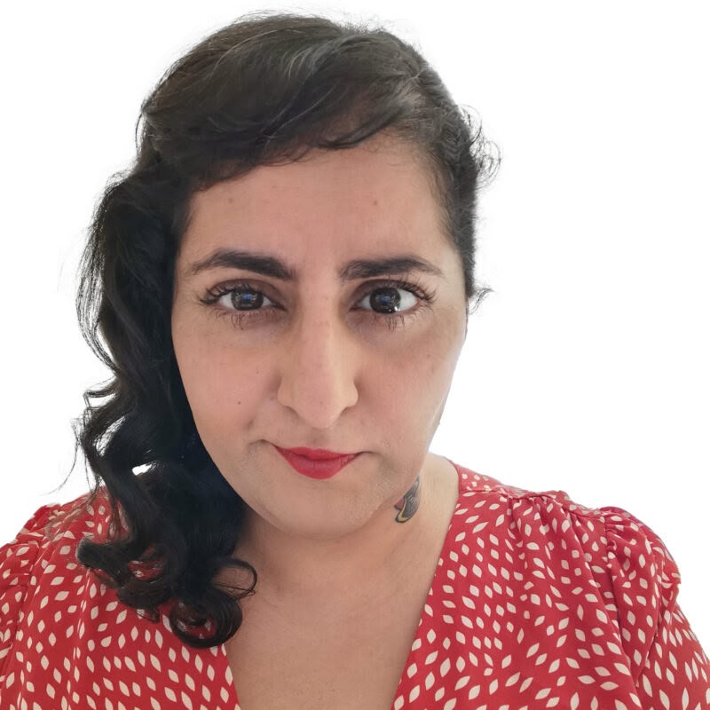 A portrait photo of a brown skinned femme person with long black curly hair. They wear red lipstick and a red and white polka dot dress.