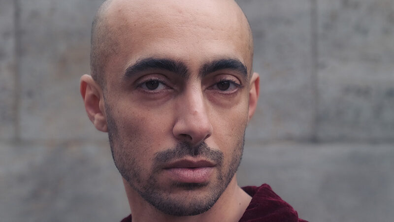 Portrait photo of Madi Awadalla, a brown skinned person with a shaved head and stubble, stood against a stone wall. They wear a red velvet shirt and are looking at the camera.