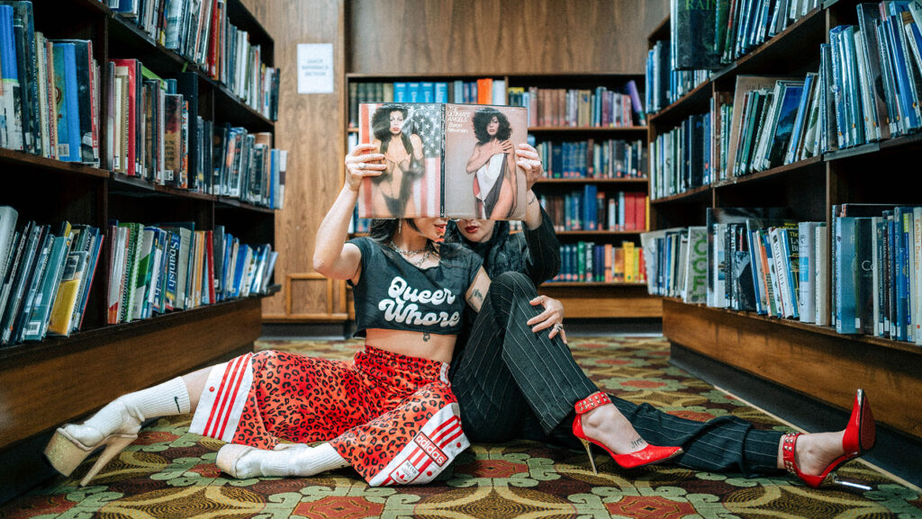 Two glamorous people in high heels sitting on a highly patterned carpet in the Mitchell Library are holding up a copy of “Ultimate Angels” which covers their faces.