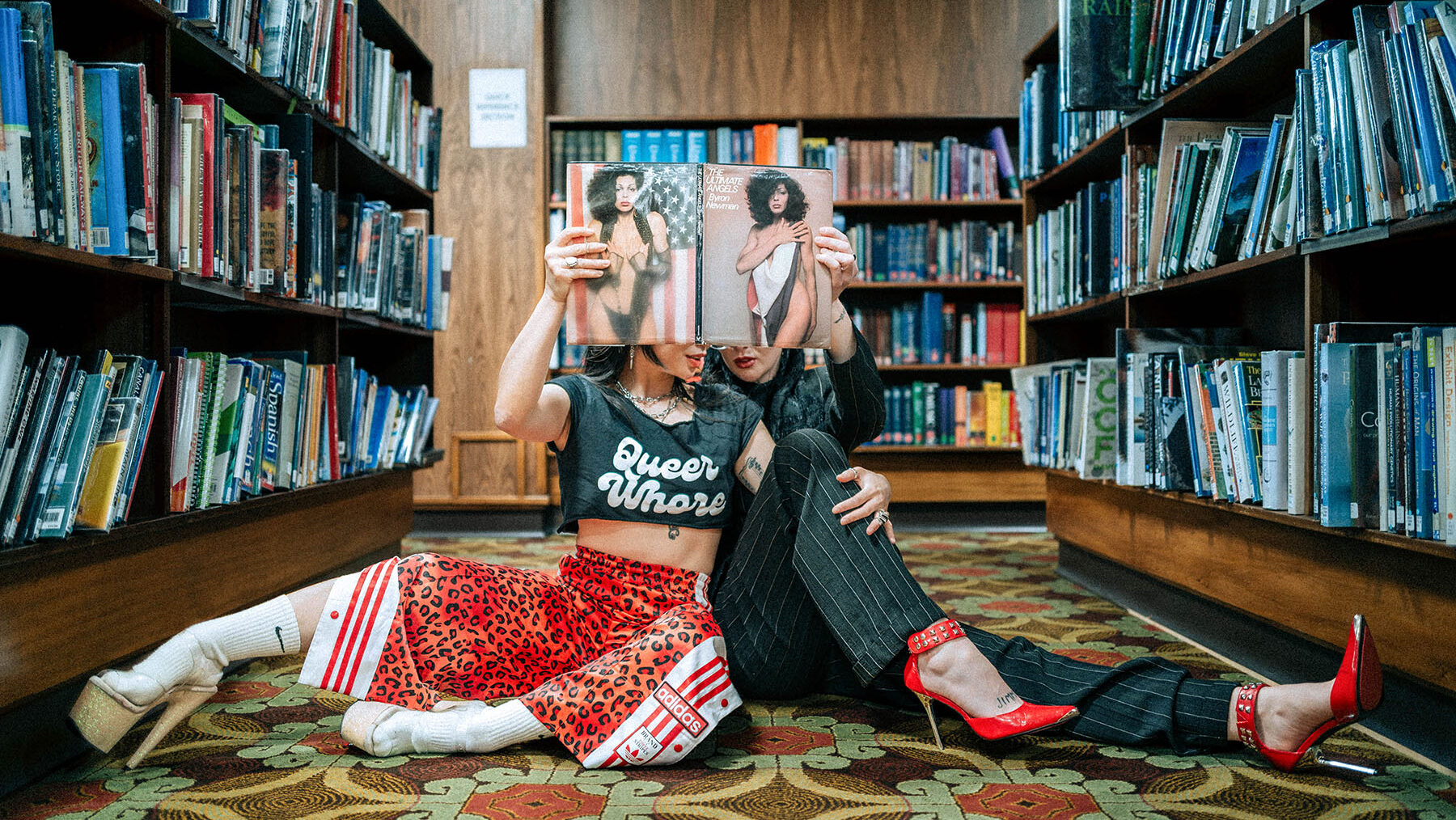 Two glamorous people in high heels sitting on a highly patterned carpet in the Mitchell Library are holding up a copy of “Ultimate Angels” which covers their faces.
