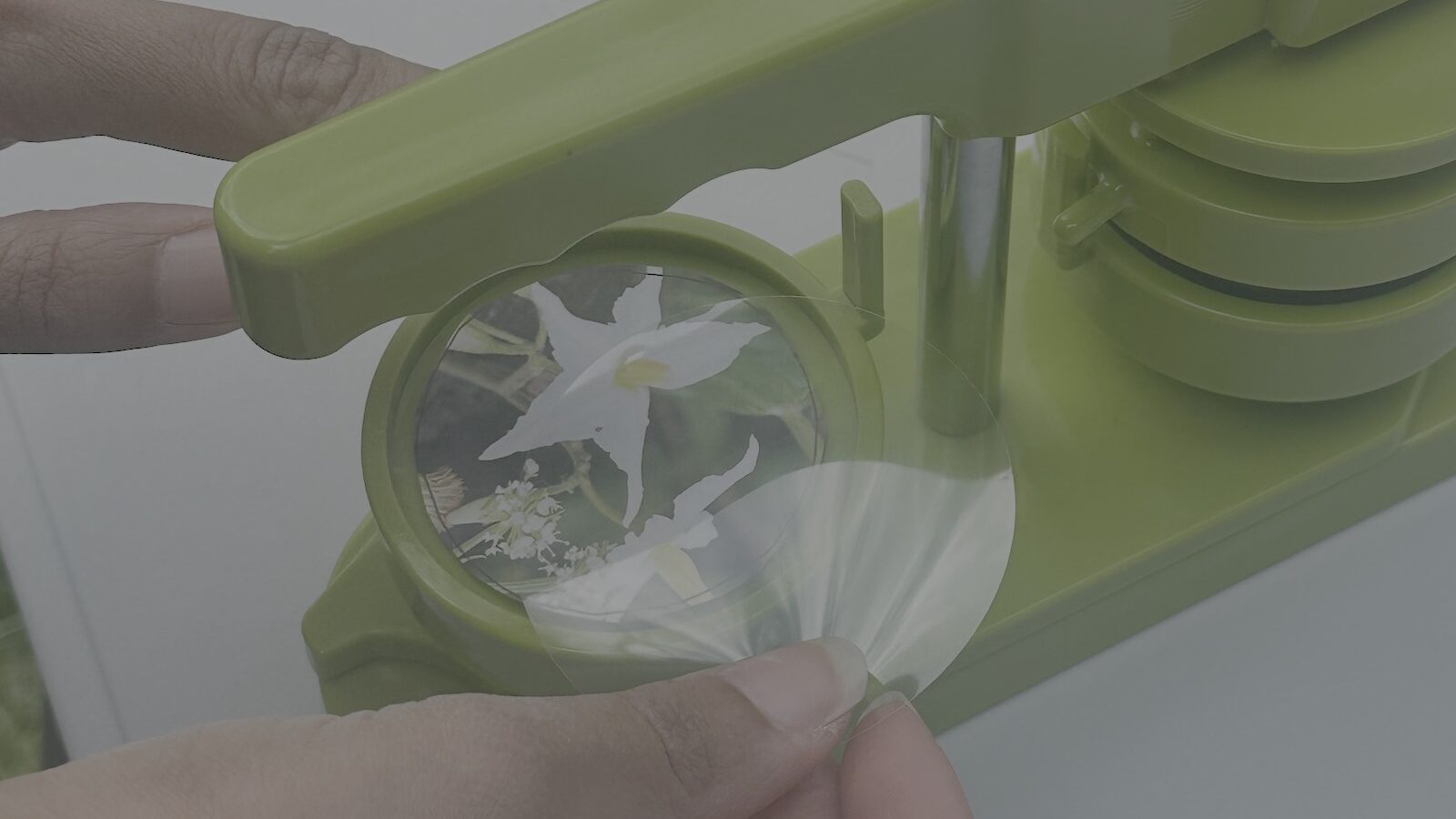 Image of green badge making machine, containing a badge with a white flower in it and two brown hands with delicate tattoos adjusting materials.