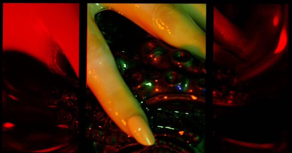 Photograph of red light glowing upon a shiny wet white hand, with long neatly manicured pale nails, sliding atop some dark twisted tentacles.