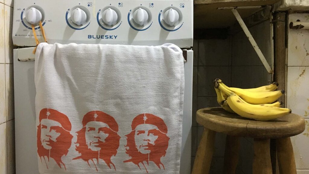 A photo of a dishcloth with three stencils of Che Guevara screen printed on it. It hangs on the handle of an electric oven in a kitchen, beside it a hand of bananas rests on a wooden stool.