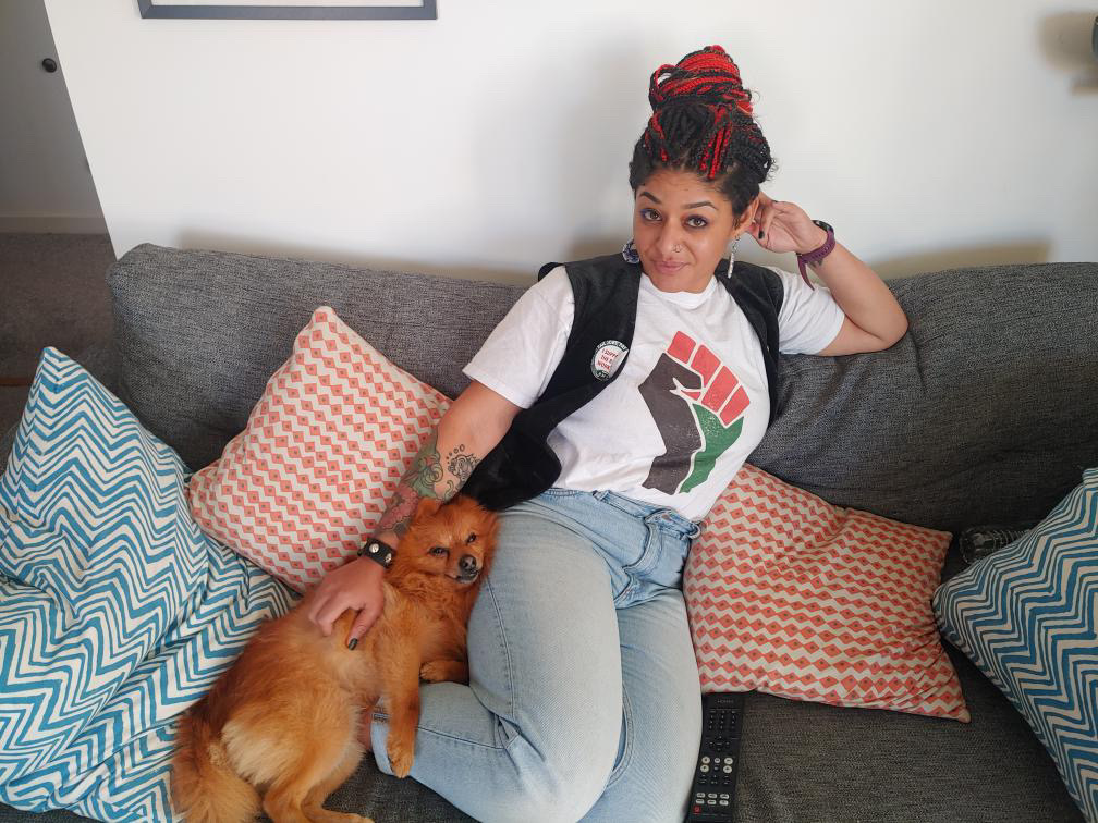 layla a black woman with brown skin, eyes, and black and red hair styled in long braids which are tied up. She is sat reclining on a sofa with a small red dog lying beside her. She wears a white T-shirt with a "raised fist" design in the Palestine flag colours, a black waistcoat with badges and pins on it, and pale blue skinny jeans.