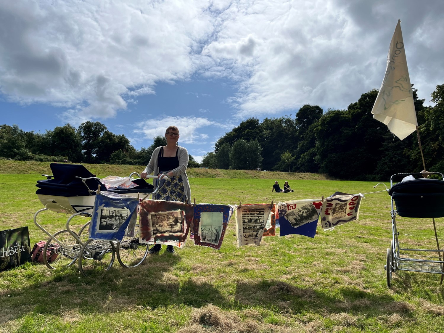 An older woman with short grey hair and light skin, she wears a grey cardigan and colourful skirt. She is stood in a wide grassy field on a sunny day. She is posed pushing an old fashioned baby's buggy, and in front of her colourful pieces of fabric are hung on a washing line.