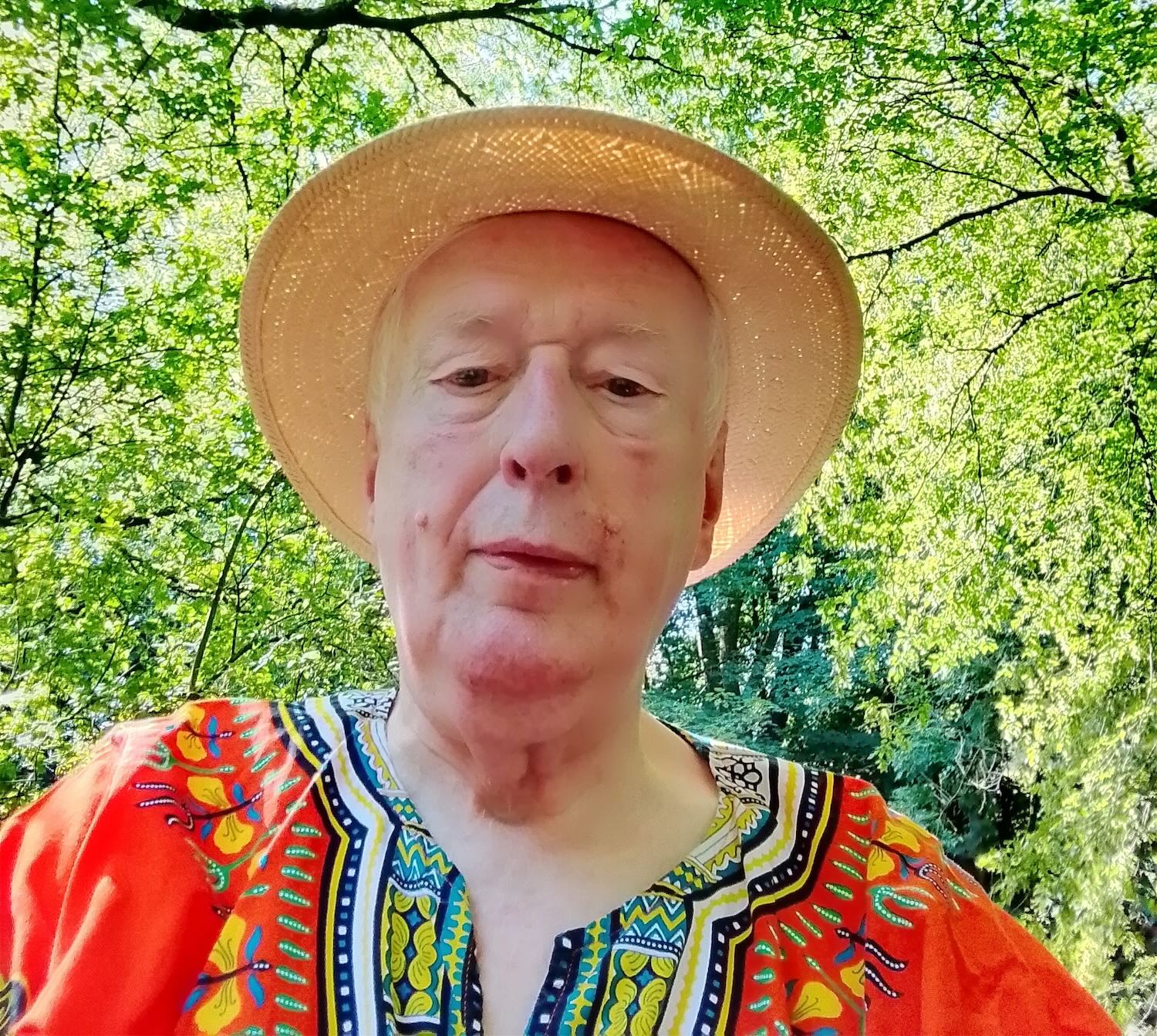 An older white man with light skin, hazel eyes, and white hair. He is wearing a straw sun hat and colourful patternred shirt which appears to be batik dyed. He is stood outside on a sunny day with lush green trees behind him.