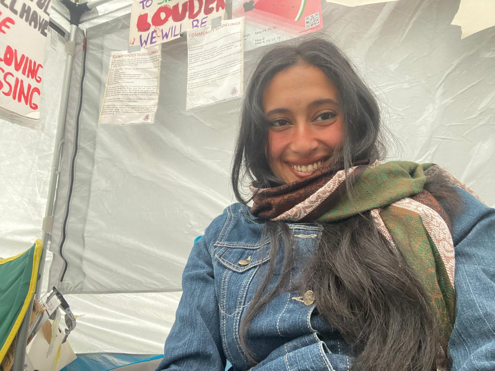 A selfie photo of Hajar, a woman with brown skin, long dark brown hair, and brown eyes. She wears a blue denim coat and is bundled in a warm looking green and brown scarf. She is sat within a white tent which has hand written signs taped to the walls. She is smiling widely and making eye contact with the camera lens.
