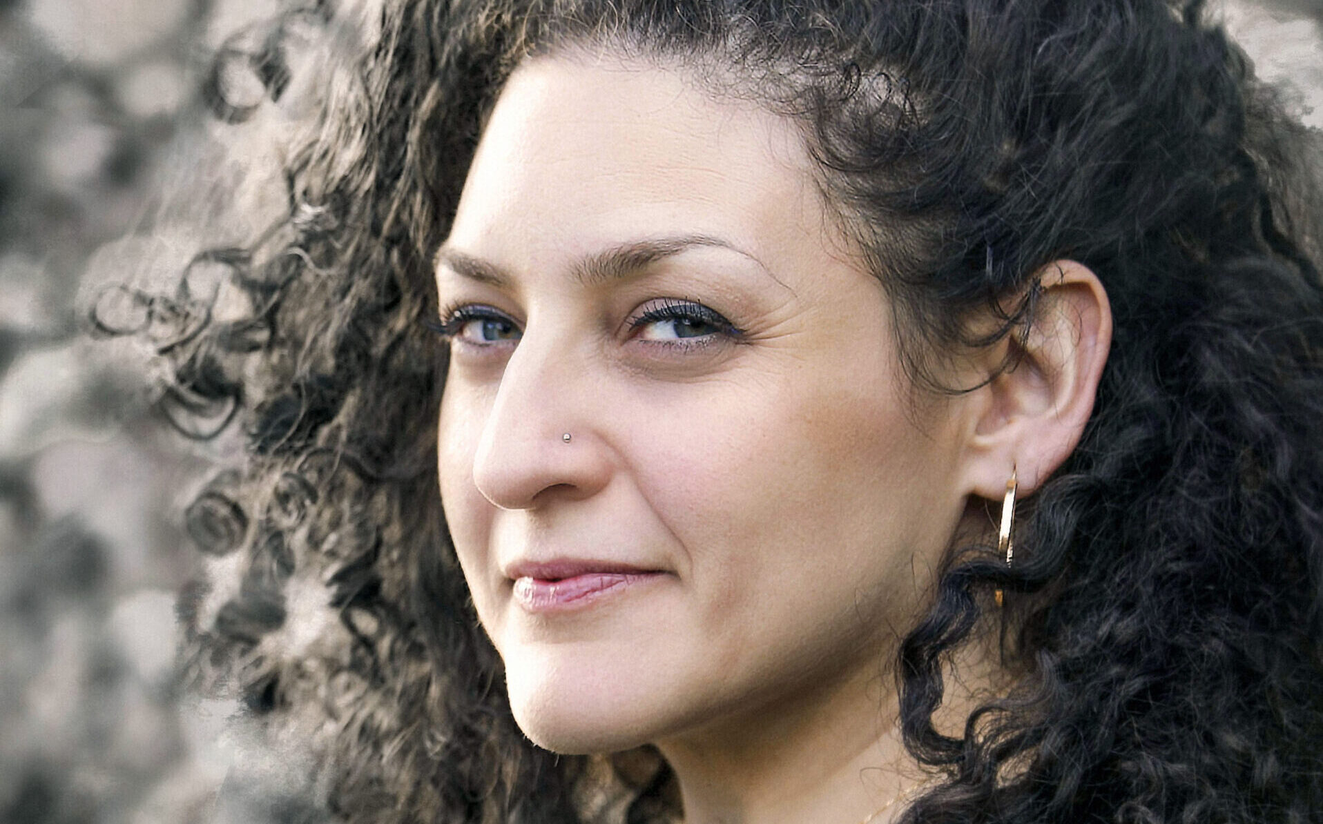 A photo of Lara Sheehi, a woman with long dark brown curly hair, olive skin, and dark hazel eyes. She wears a black vest, gold hoop earring, and a stud nose piercing.
