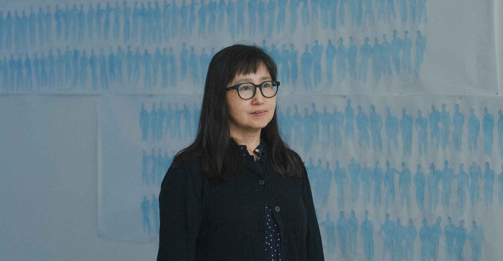 Don Mee Choi a well groomed Korean woman with medium long hair that ends on her collar bone. She has dark brown hair with a few greys throughout. She is wearing a black cardigan and a dotted white and blue shirt. She is stood in front of a series of blue ink drawings of shilouetted people on paper.