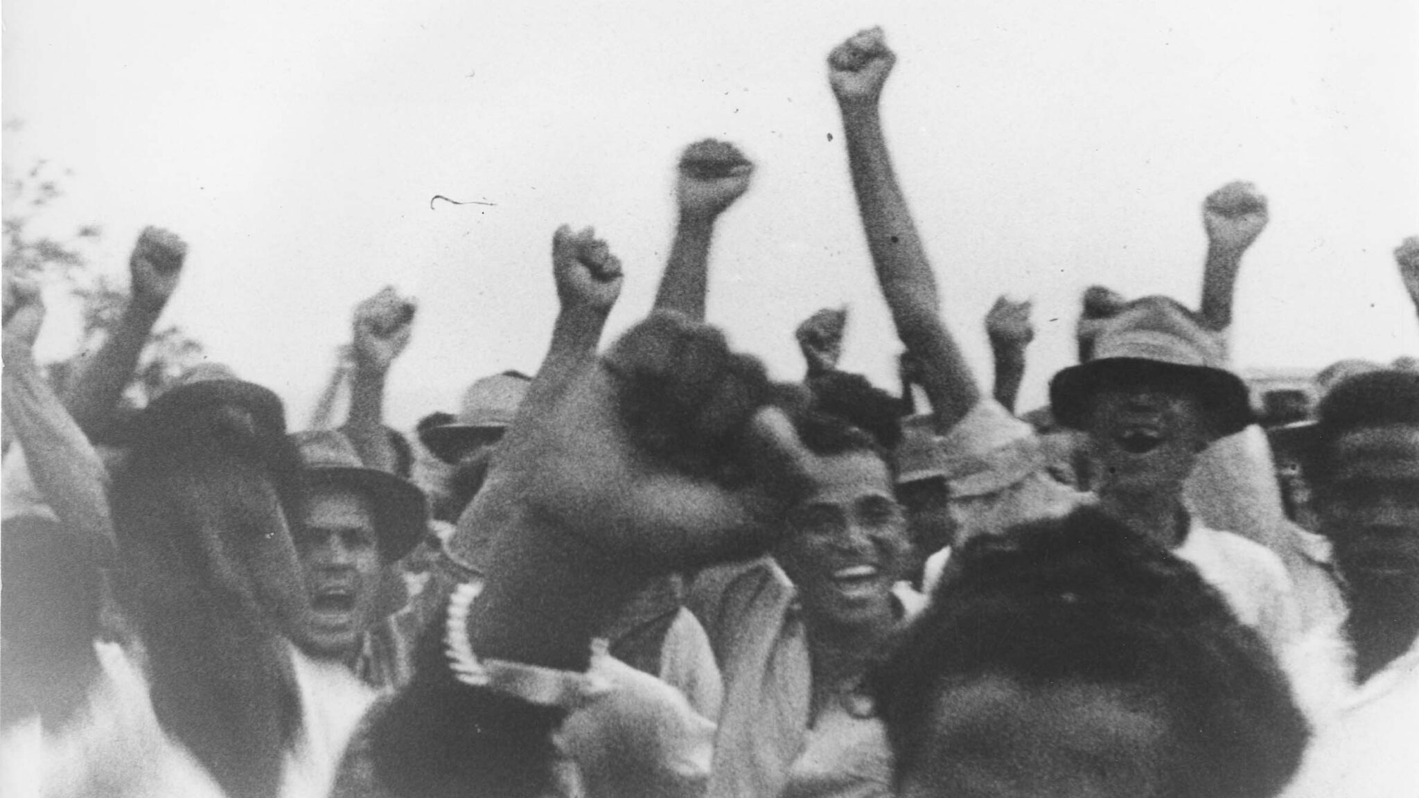 A grainy black and white film still from the 60s: Depicting a group of Reunionese activists. The crowd is mainly men, dressed in loose shirts and sun hats. They hold their fists in the air in celebration and are smiling.