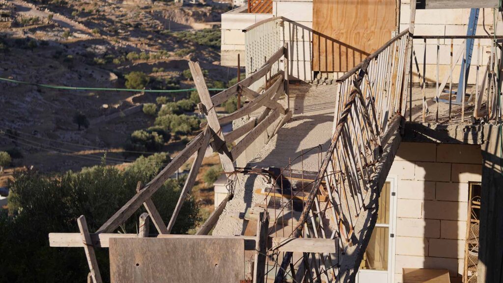 An exterior photo of a portion of a damaged building. The destroyed stairway has been re-constructed with concrete, scrap metal, and salvaged wood. And the door has been replaced with plywood. In the background there is a rolling hill covered in olive trees, shrubland, and other damaged buildings.