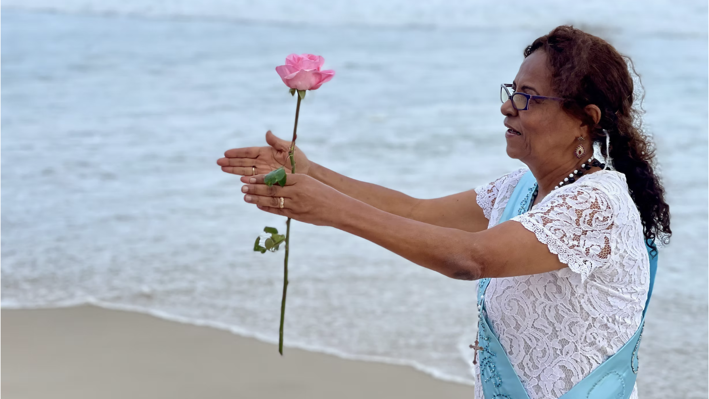 Leda stood at a beach with the sea behind her. Her hands are reaching out ceremonially, with a pink rose held between her palms. She wears a white floral dress with a baby blue sash, a gold decorative cross hangs on one side.