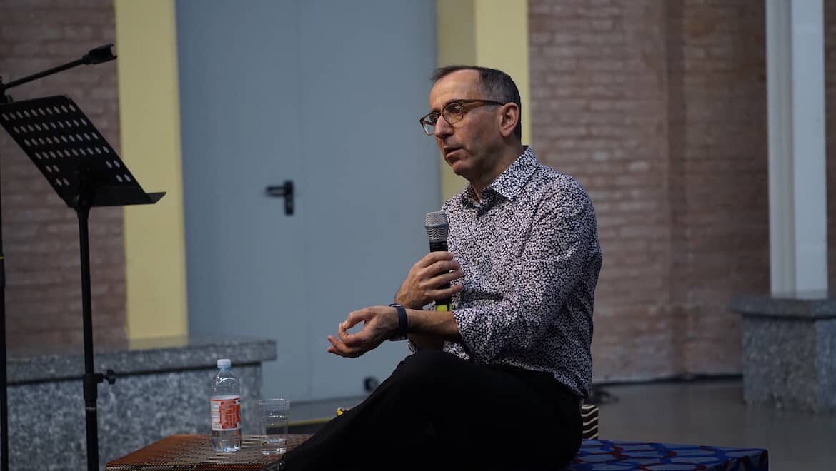A man wearing a patterned formal shirt and black trousers. He has light skin and dark hair which is greying at the temple. In one hand he holds a microphone in the other he gestures openly, he appears to be thoughtfully responding to a question.