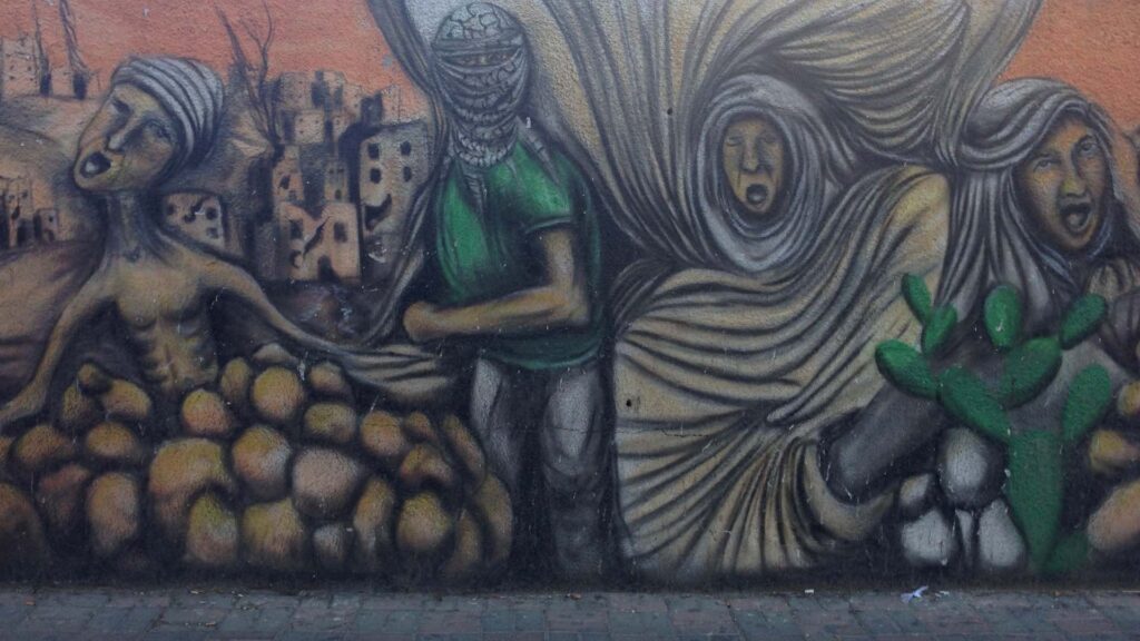 A mural on a brick wall. The painting shows four people: One is shirtless and appears to be half buried under rubble, another’s face is covered by a keffiyeh and holds a strong defiant pose, the other two are wearing hijab and scream in anguish. Behind them a landscape of destroyed building can be seen.