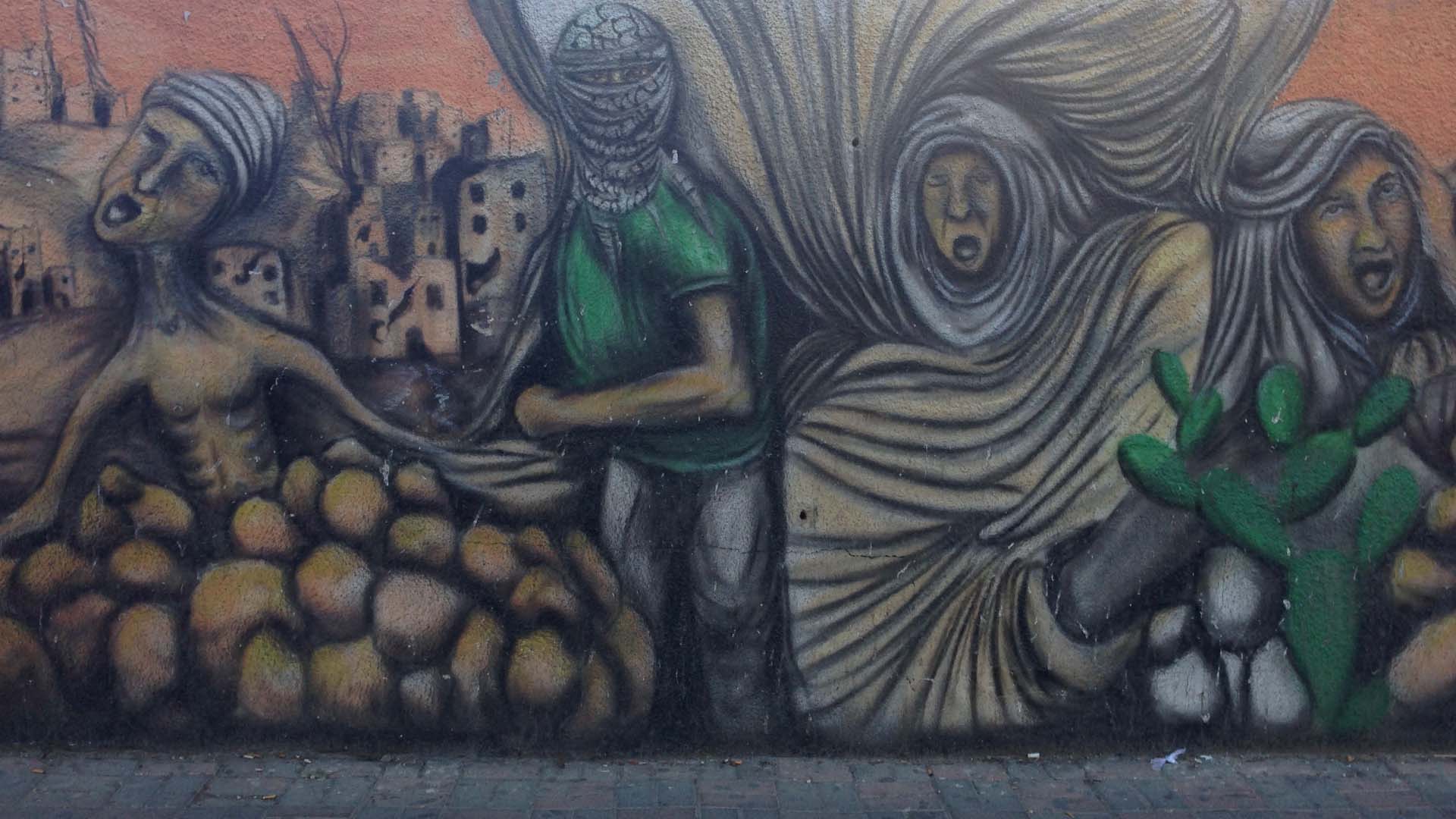 A mural on a brick wall. The painting shows four people: One is shirtless and appears to be half buried under rubble, another’s face is covered by a keffiyeh and holds a strong defiant pose, the other two are wearing hijab and scream in anguish. Behind them a landscape of destroyed building can be seen.