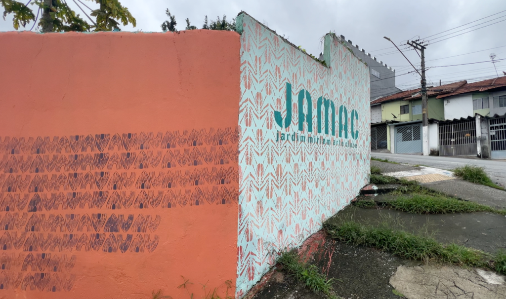 A photo of an external wall in a deprived looking urban area. One side of the wall is painted orange with a repeating arrow pattern in black, around the corner it is painted white with the pattern in orange. The text “JAMAC Jardim Miriam Arte Clube” appears in green stencilled letters on the white wall.