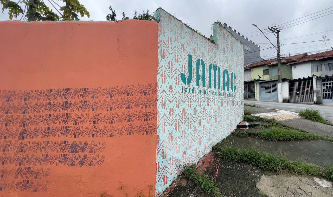 A photo of an external wall in a deprived looking urban area. One side of the wall is painted orange with a repeating arrow pattern in black, around the corner it is painted white with the pattern in orange. The text “JAMAC Jardim Miriam Arte Clube” appears in green stencilled letters on the white wall.
