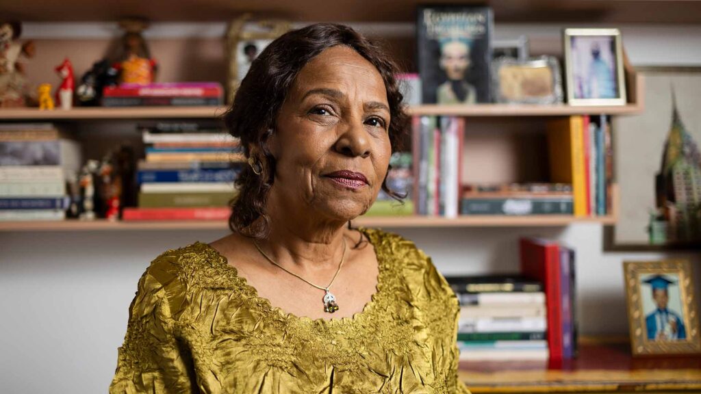 A black woman in her 60s with brown skin, hair and eyes. She wears a ruffled yellow satin dress, red lipstick, gold earrings and a silver pendant necklace. Behind her are carefully curated shelves covered in books, trinkets, and photographs—possibly of friends or family members.