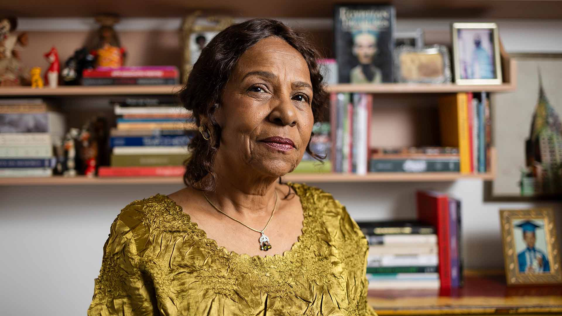 A black woman in her 60s with brown skin, hair and eyes. She wears a ruffled yellow satin dress, red lipstick, gold earrings and a silver pendant necklace. Behind her are carefully curated shelves covered in books, trinkets, and photographs—possibly of friends or family members.