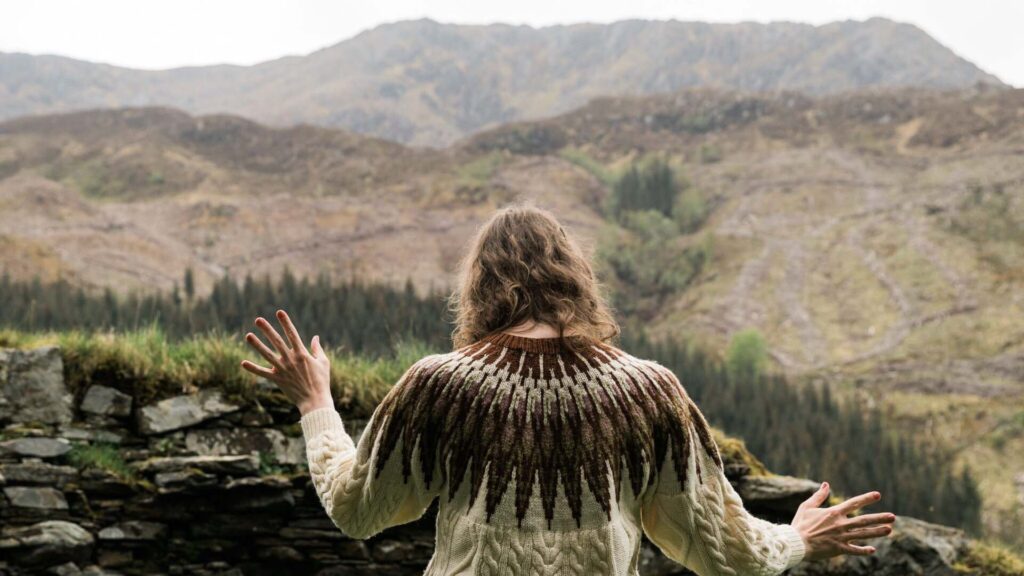 A photo of a woman facing away from the camera, she has pale skin and wavy light brown hair and wears a knitted jumper. The background is a wide expanse of mountains and hills, her hands are posed in ritualistic or ceremonial manner.
