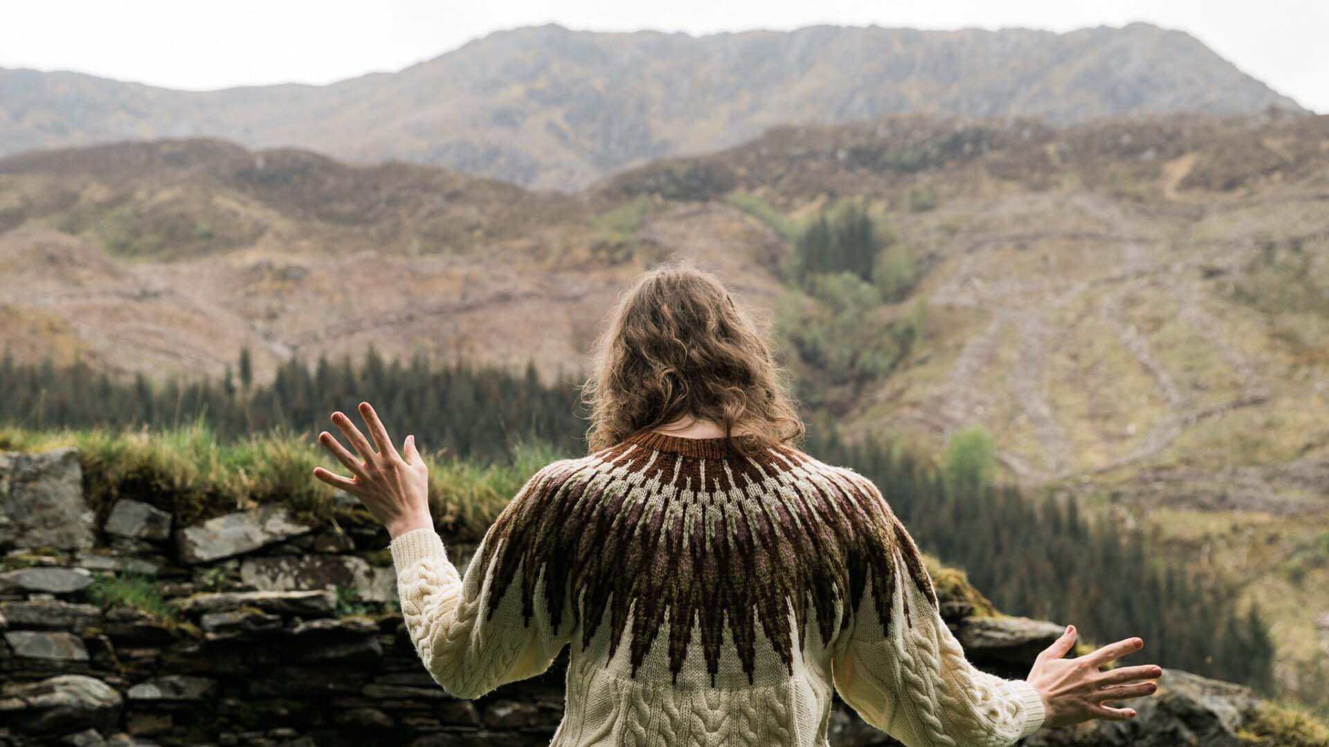 A photo of a woman facing away from the camera, she has pale skin and wavy light brown hair and wears a knitted jumper. The background is a wide expanse of mountains and hills, her hands are posed in ritualistic or ceremonial manner.