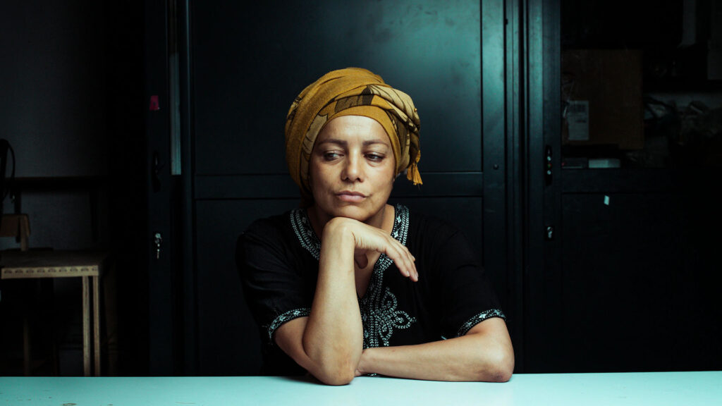 A black woman with brown skin sat in a formal looking space. There black wood panel walls behind her and she leans her elbow on a stark white table. She is resting her chin on her wrist pensively and looking away from the camera.