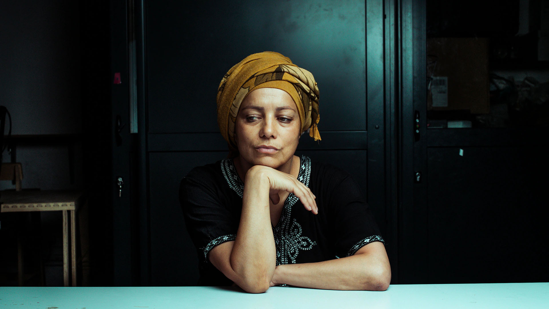 A black woman with brown skin sat in a formal looking space. There black wood panel walls behind her and she leans her elbow on a stark white table. She is resting her chin on her wrist pensively and looking away from the camera.