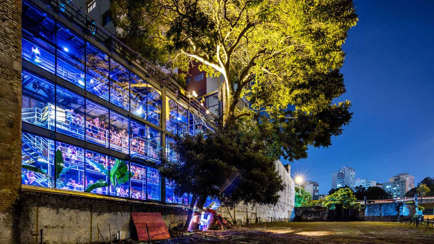 An external shot of the Teatro Oficina at night. A tall narrow building with tall windows that span across multiple floors. The interior is filled by scaffolding, which serves as seating. It is lit in blues and reds. Besides the building a tall tree edges next to the building and an empty lot sits beside it.