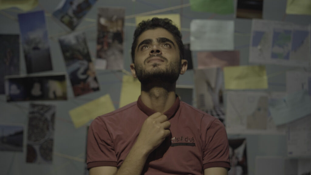 A head and shoulders photo man with brown skin and dark brown eyes and hair. He has thick eyebrows and a short groomed beared and wears a red polo shirt. He is oriented to the camera with his head upturned, his other hand tugs his collar in what could be anxiety. Behind him is a web of images, articles, and papers connected by strings.