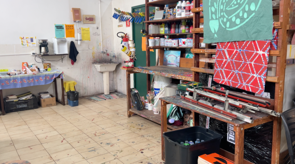 An image of a busy screen printing studio, ink splatters the tiled floors, printed fabrics hang on the walls, and a messy sink is visible in the background.