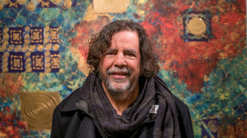 A portrait of a middle aged man with light tanned skinned, a short salt and pepper beard, brown wavy hair, and dark eyes. He wears all black including a scarf and stares into the camera. He's in front of a brightly coloured painting full of abstract pattern in gold, green, red and yellow.