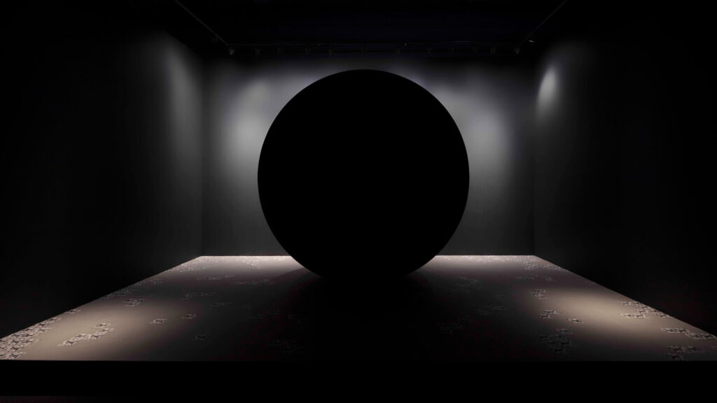 A pitch black sphere sat in a dark space. It spot lit by lights and is so black it appears to be a void in space.