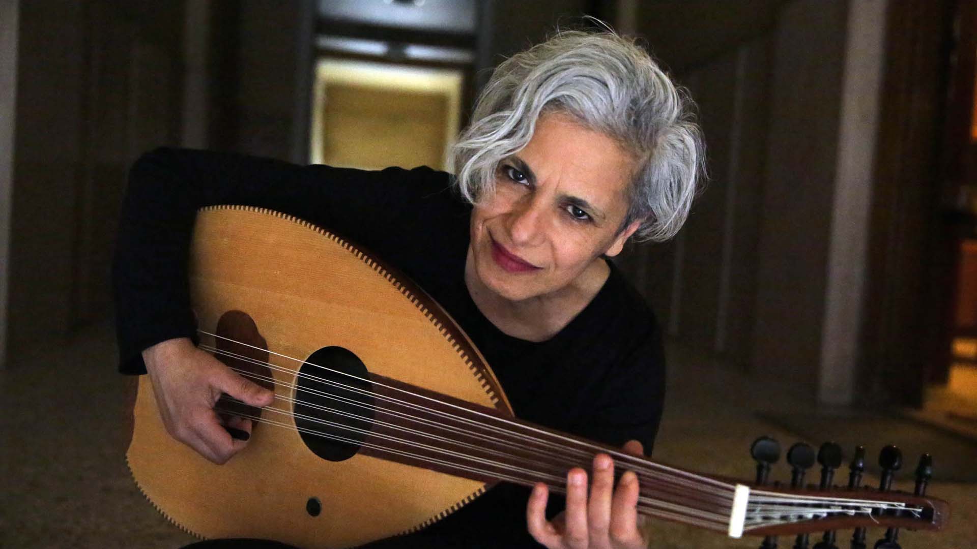 A woman with stylish short grey hair, olive skin sat down holding an Oud and leaning towards the camera.