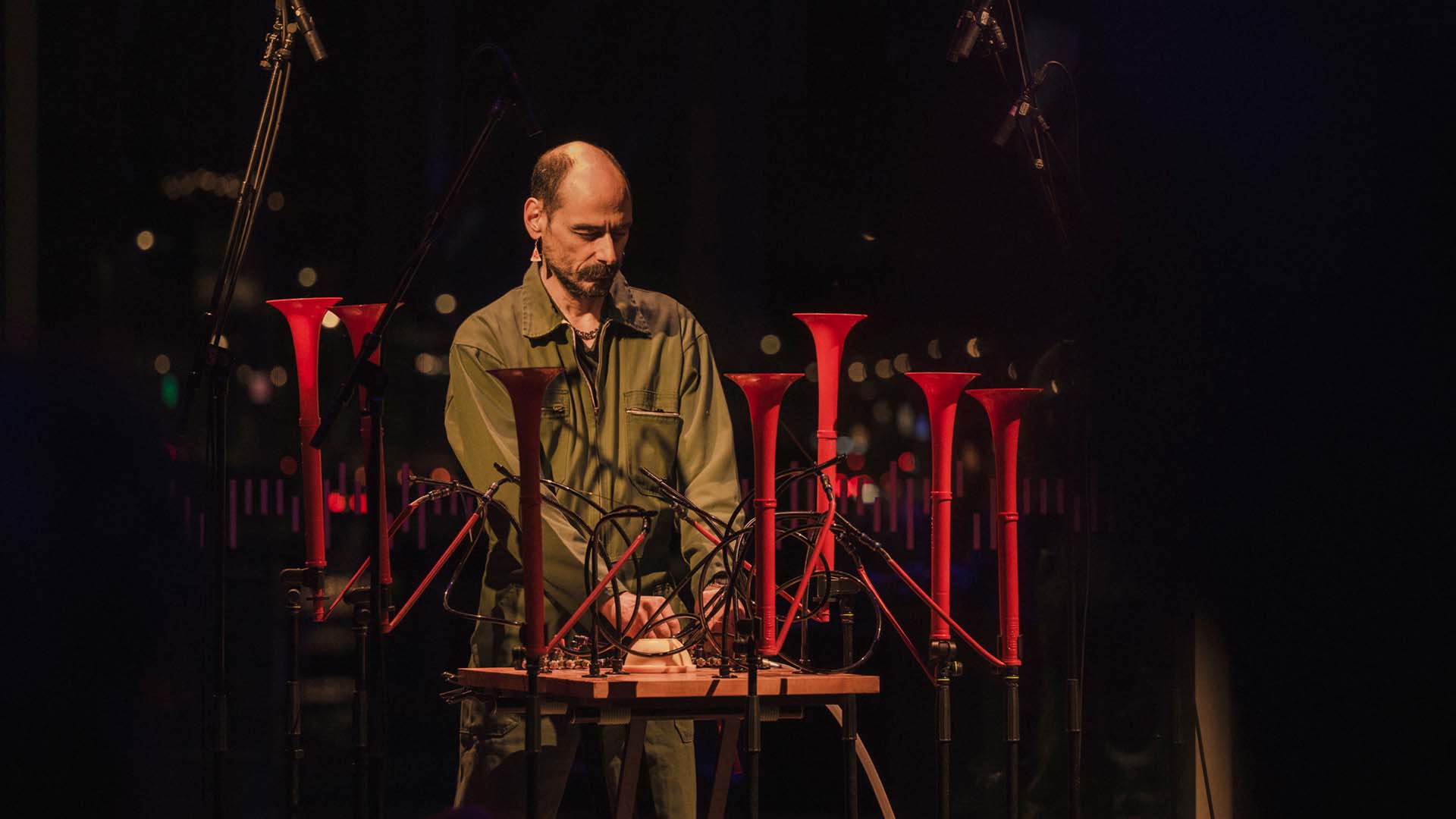 A man with a thick moustache wearing a khaki boilersuit and watermelon earing. He plays a strange instrument made of 7 vertical red trumpets connected via pipes to a wooden control panel and compressed air units.