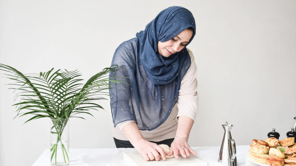 A woman with light skin and dark hair wearing a blue sequinned hijab. She is preparing a pastry. The photo is clean and composed.