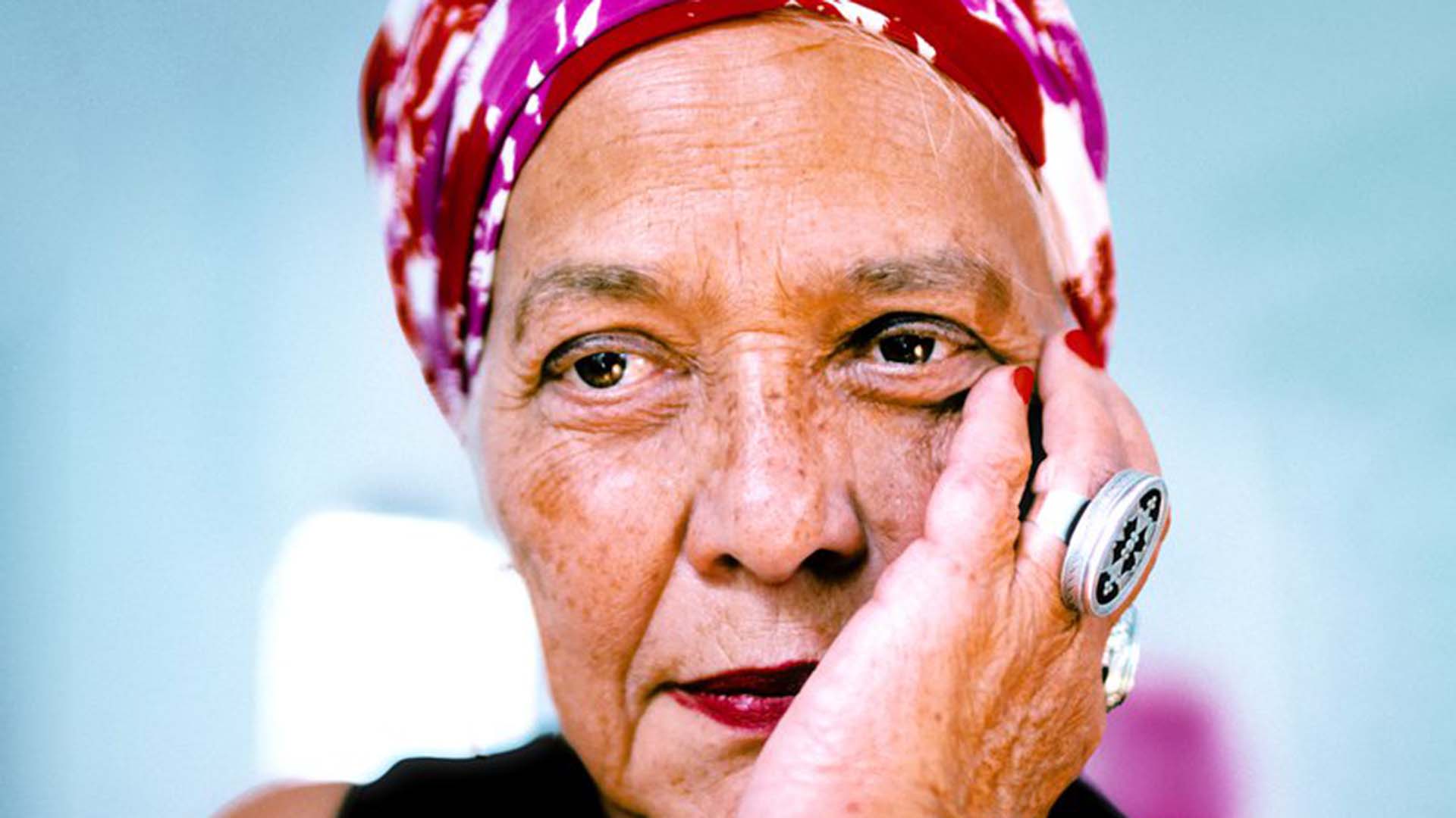 A portrait photo of Françoise Vergès, an older woman with light olive skin, freckles, and brown eyes. She is leaning on her hand and wears a pink floral headscarf, red lipstick, and chunky jewellery.