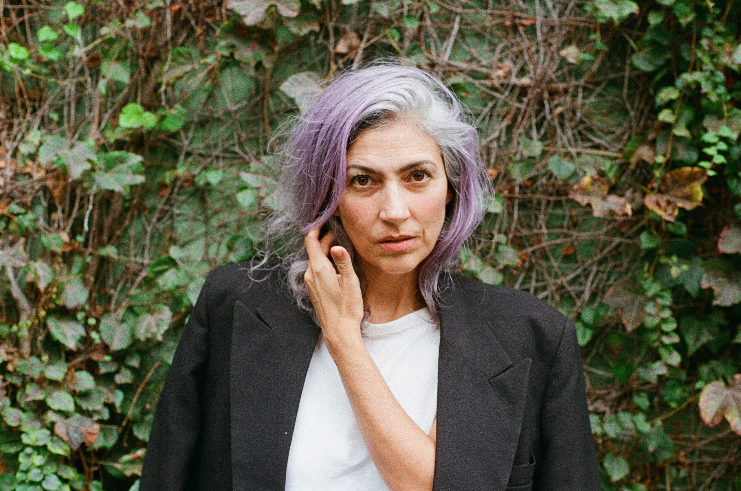 A slim middle aged woman with olive skin, grey and purple hair. She wears a black blazer over a white t-shirt and is stood in front of a mess of ivy.