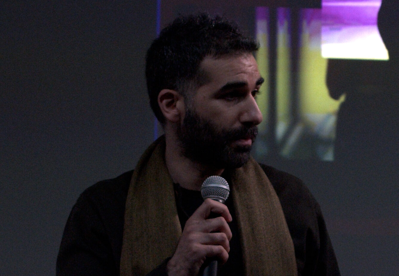 Nasser Abourahme speaks into a microphone with abstract projected images behind.