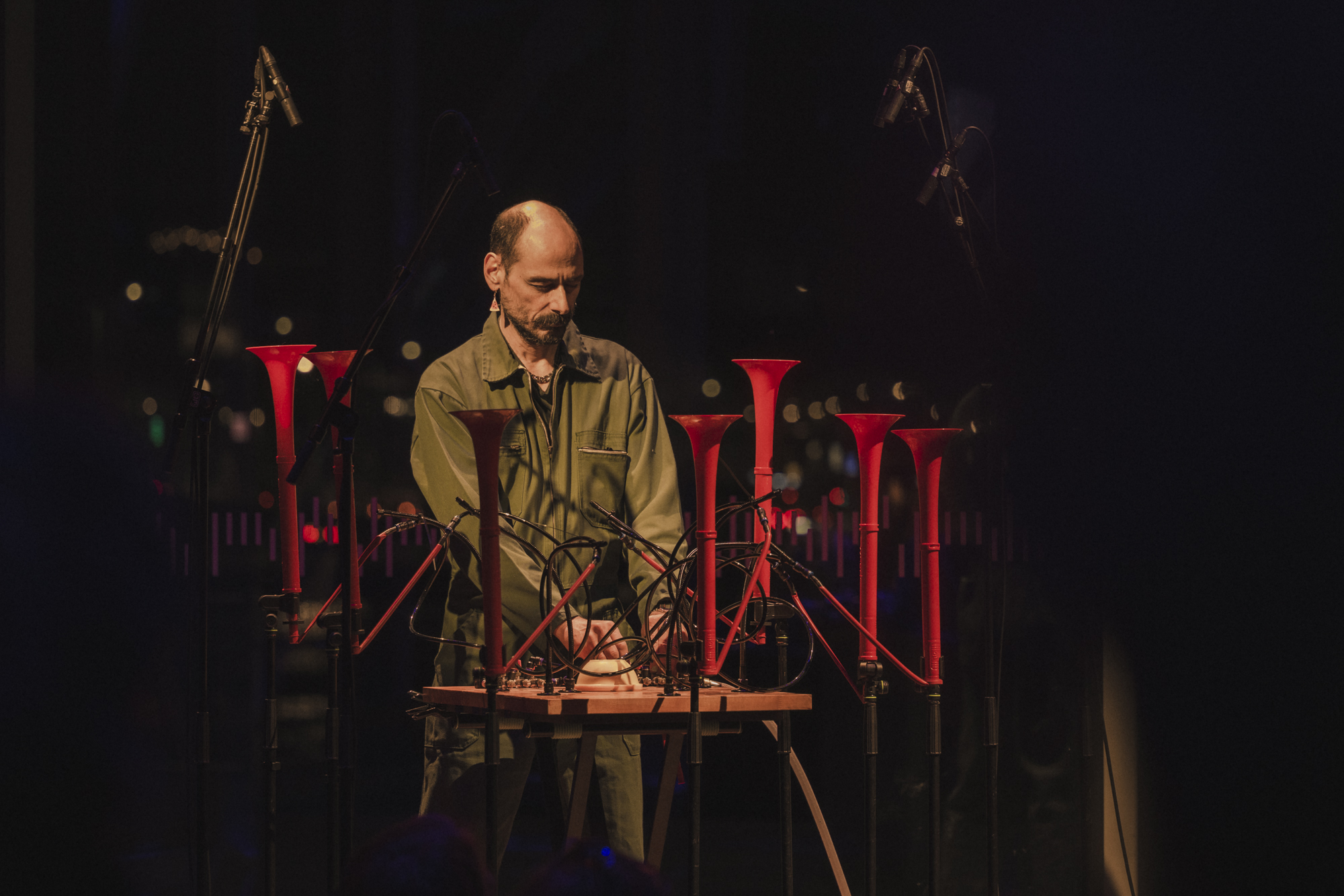 mazen kerb performing, stood behind an unusual musical instrument made up of mixing desk that's attached to 7 red trumpet-like horns facing upward.