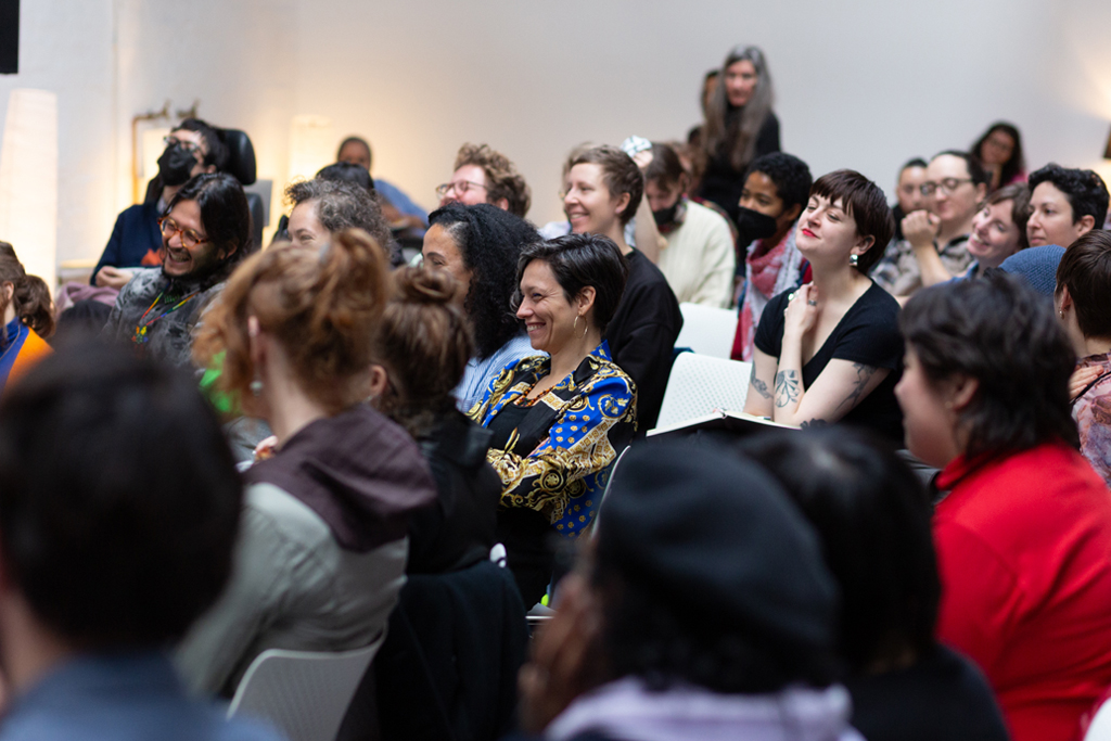 An audience smiling, looking and listening attentively in a bright white room.